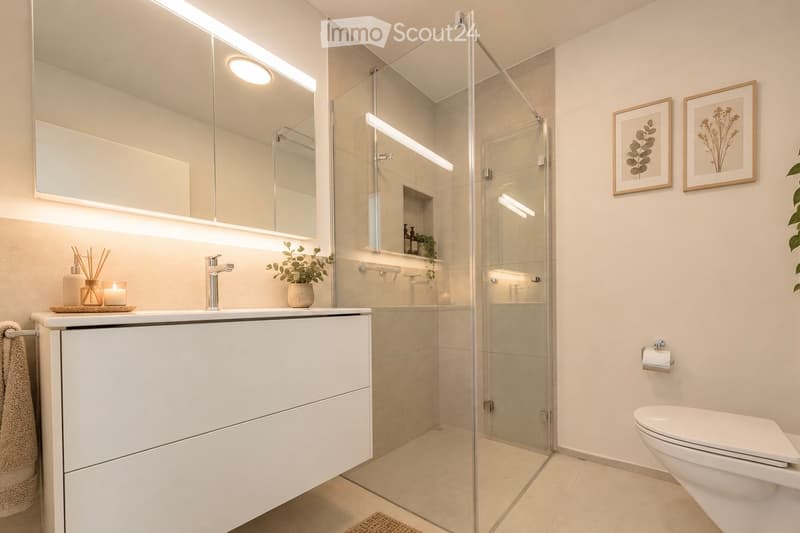 Modern bathroom with a white vanity, sink, mirror, and a glass-enclosed shower. Includes a toilet, towel, small plant, and framed pictures on the wall.