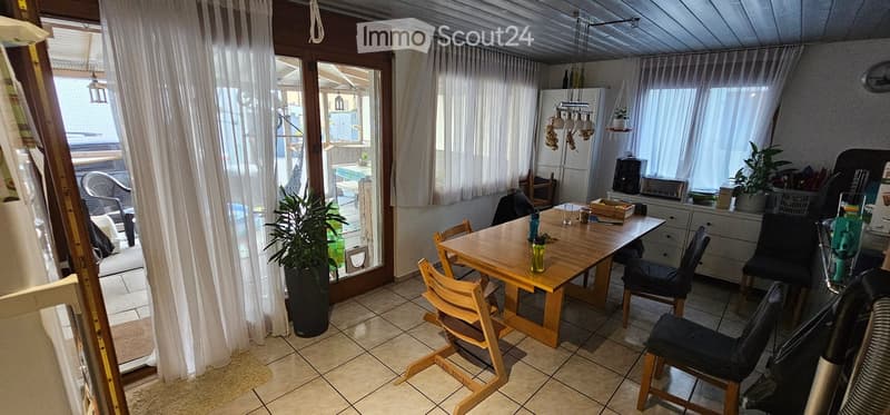 A well-lit dining area with a wooden table and chairs, a refrigerator, and a curtained window leading to a patio. There are plants and a small cabinet visible.
