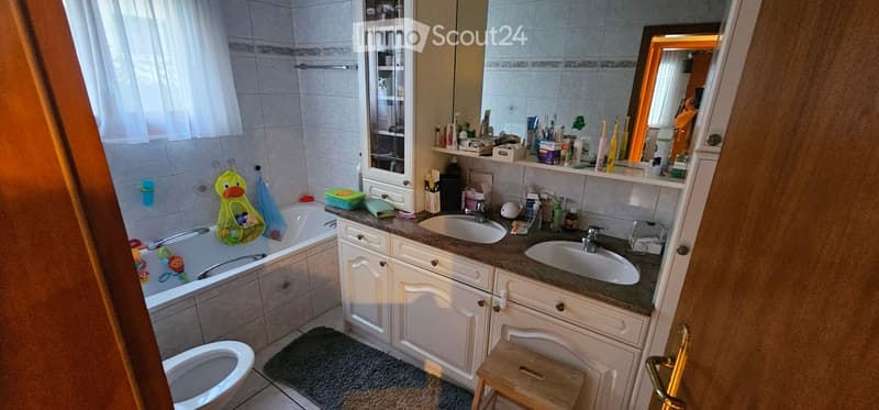 Bathroom with a bathtub, double sink, mirror, and various bathroom accessories and products. Includes a small stool and a toilet.