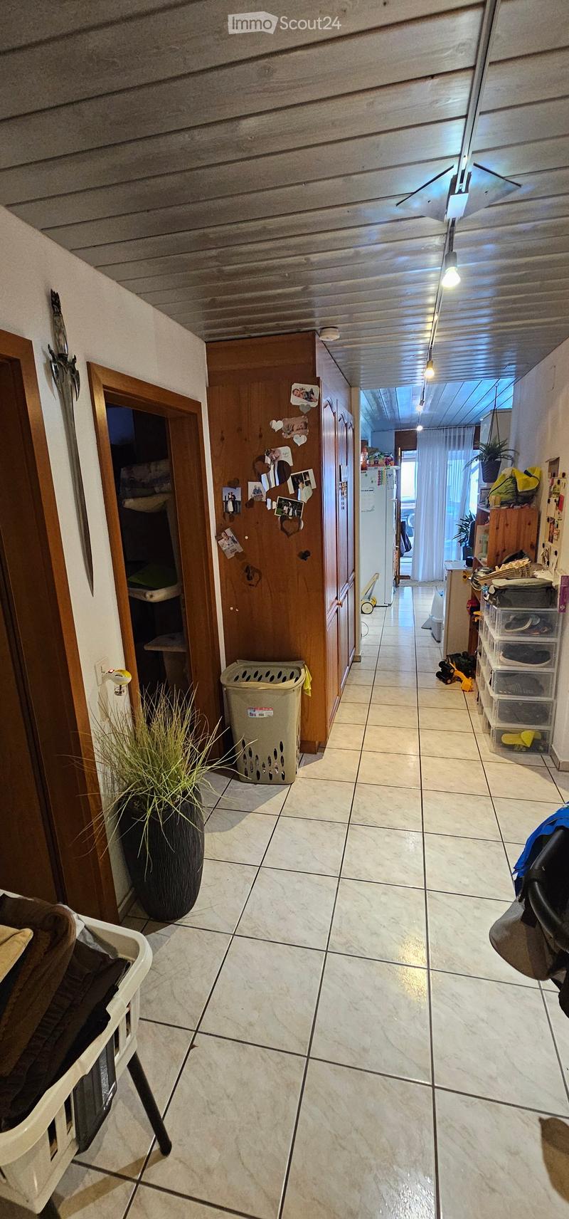 Hallway with tiled floor, wooden doors, and visible storage items and a laundry basket. The hallway leads to a kitchen area with a refrigerator and various household items.
