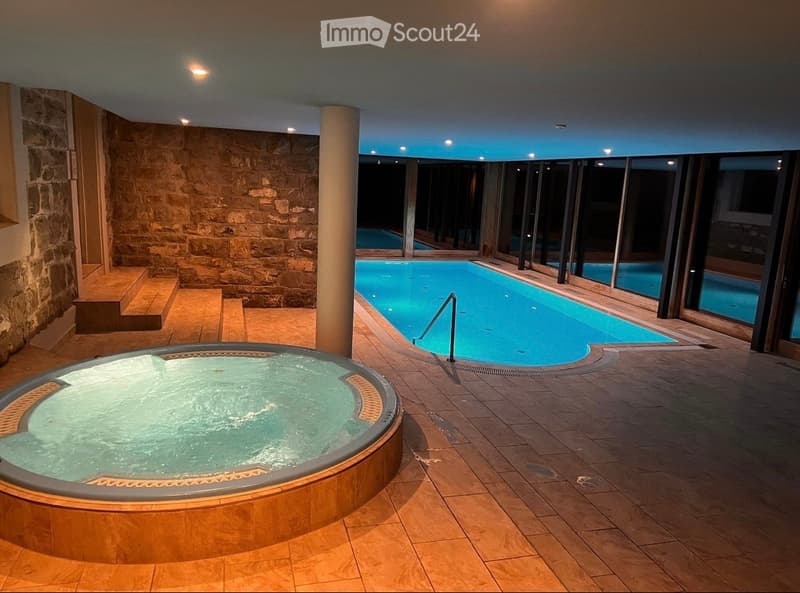 Indoor pool area with a hot tub, swimming pool, stone walls, and large windows offering a view of the outdoor space.