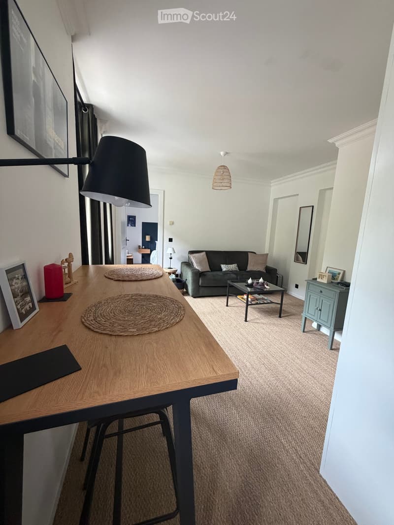 Modern living room with a grey sofa, coffee table, wooden side table, and a hanging lamp. The space features light-colored walls and carpeted flooring.