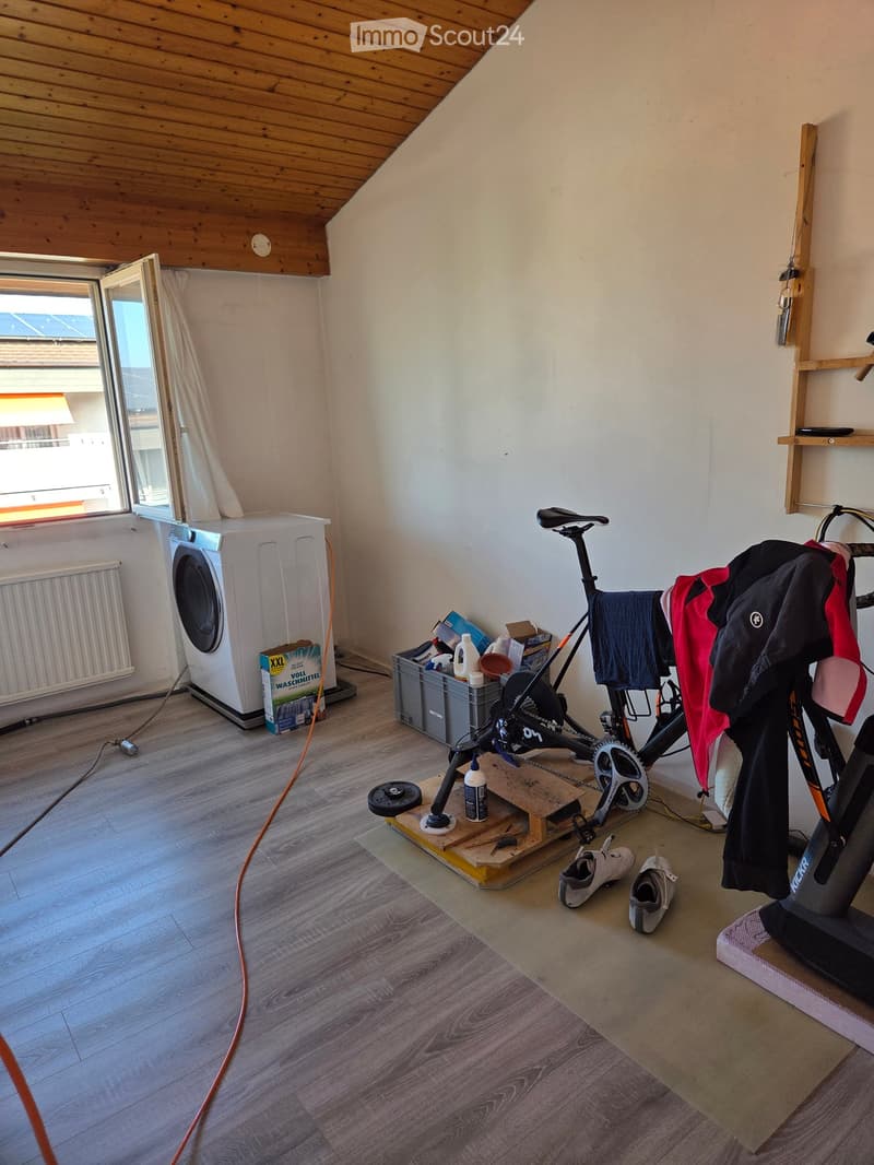 Room with wooden flooring, slanted wooden ceiling, large window, washing machine, bicycle on a stand, clothes hanging, shoes, and storage items.
