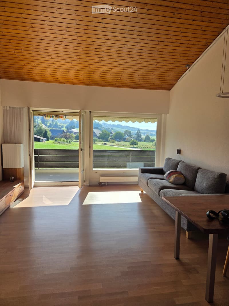 Bright living room with wooden floors, large windows offering a scenic view, a grey sofa with a colorful pillow, a coffee table, and a side lamp.