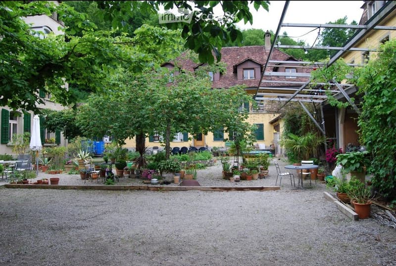 A lush garden area with gravel ground, surrounded by potted plants, trees, and outdoor furniture including tables and chairs. The background features a yellow building with green shutters and a red-tiled roof.