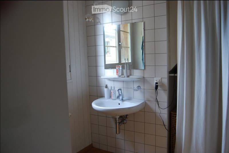 A small bathroom with a white tiled wall, a sink with a faucet, a mirror above it, and shelves holding toiletries.
