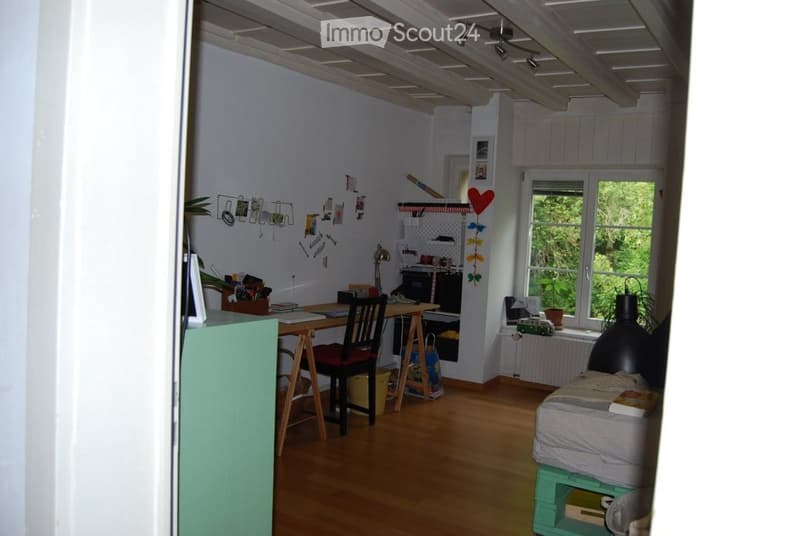 A multi-functional room with a desk, chair, bed, and various decorations including framed pictures and a plant. The room features wooden flooring and exposed wooden beams on the ceiling.