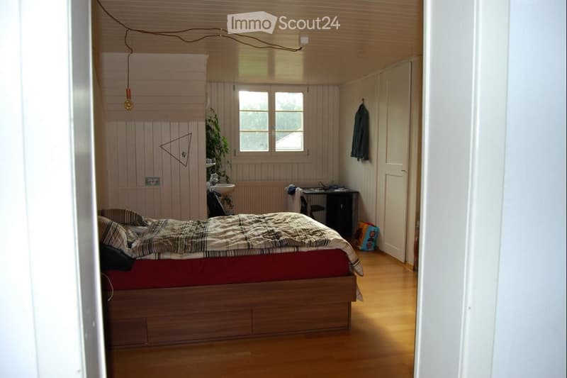 Cozy bedroom with wooden bed, desk, chair, sink, and plant. Light wooden walls and floor, with a window providing natural light.