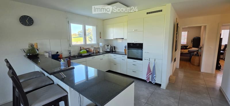 Modern kitchen with white cabinets, black countertop, bar stools, and various appliances including a sink, oven, and microwave. A clock is mounted on the wall, and a view of a green landscape is visible through the windows.