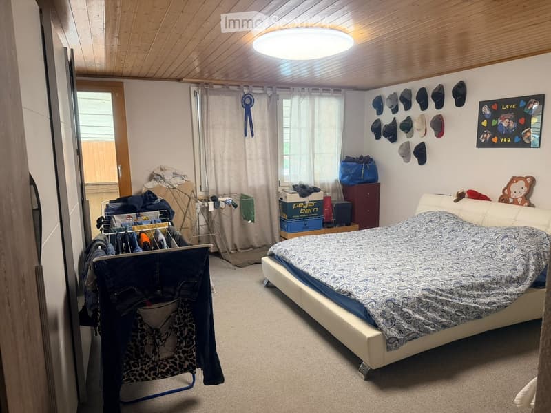 Cozy bedroom with a large bed, a small sofa, a wardrobe, and a drying rack with clothes. The walls are decorated with hats and a framed picture. The room has wooden ceilings and large windows with curtains.