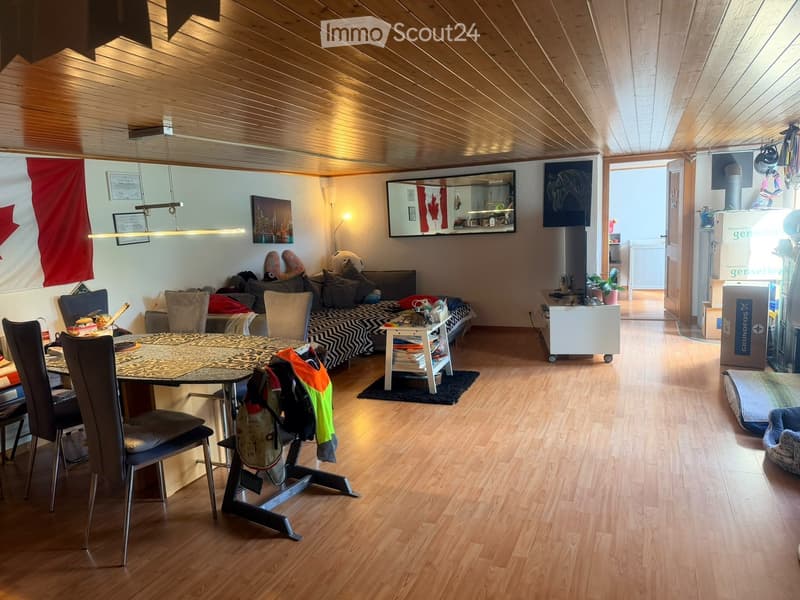A cozy living and dining area with wooden flooring, a slanted wooden ceiling, a sofa, a dining table with chairs, a small side table, a mirror, and a lamp.