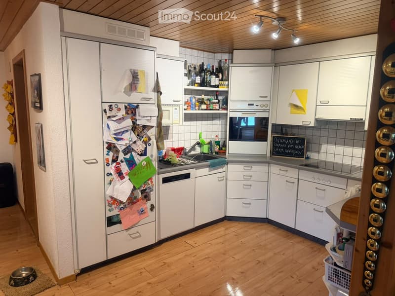 Modern kitchen with white cabinets, wooden flooring, refrigerator, oven, microwave, sink, and various bottles on shelves.