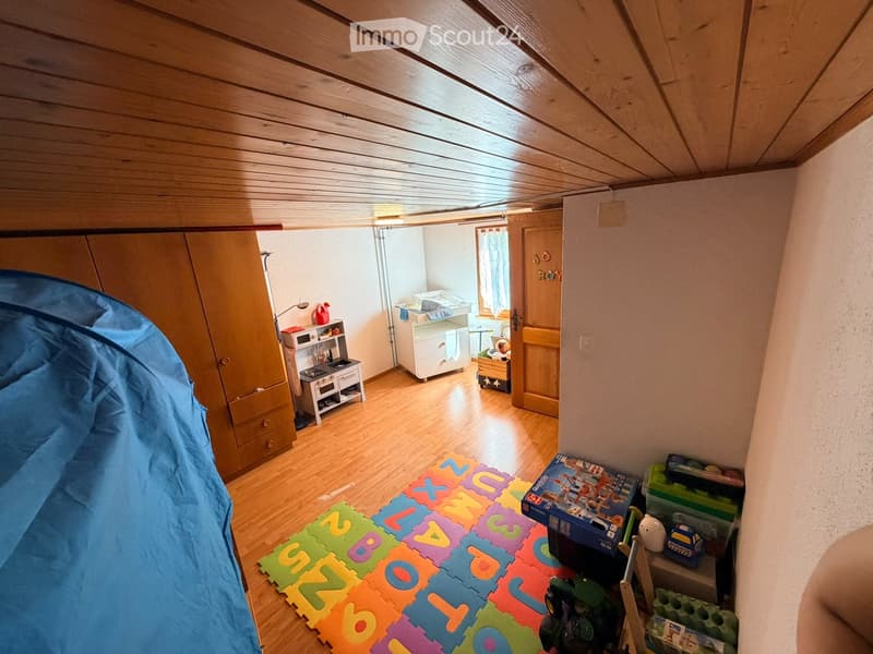 A well-lit children's room with wooden flooring, a large wardrobe, a toy stove, a crib, and colorful play mats and toys.