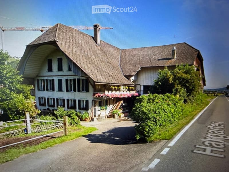 Two-story house with a steep roof, multiple windows, and a balcony adorned with flowers. The house is surrounded by greenery and a gravel driveway.