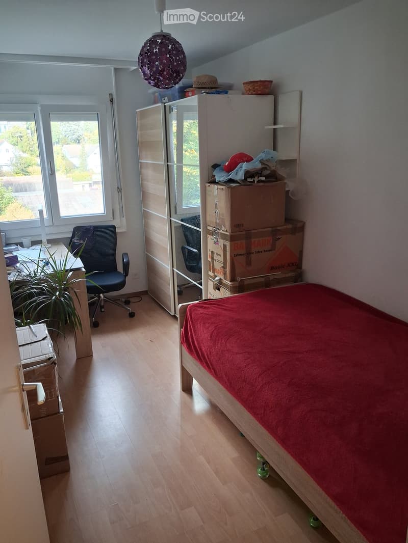 Bedroom with wooden floor, bed with red cover, desk with chair, wardrobe, and several cardboard boxes stacked around the room.