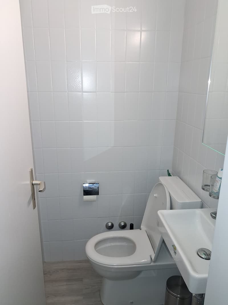White tiled bathroom with a toilet, sink, toilet paper holder, and a small trash can. A cup and bottle are placed on the sink counter.