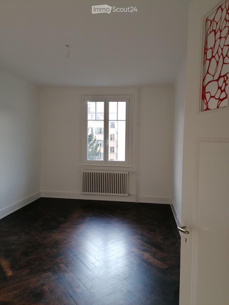 Empty room with wooden flooring, white walls, a window with white frame, and a radiator below the window.
