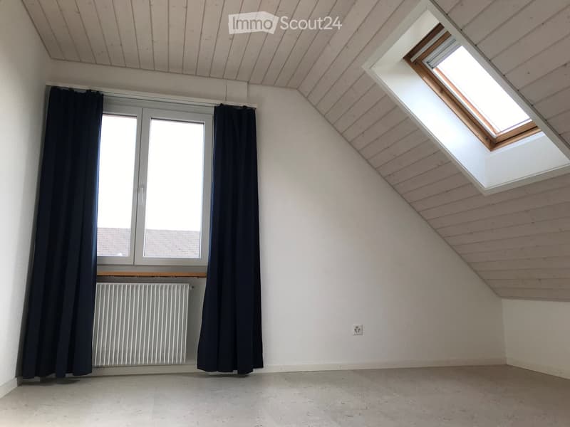 Empty attic room with wooden slanted ceiling, skylight windows, large window with dark curtains, radiator, and concrete floor.