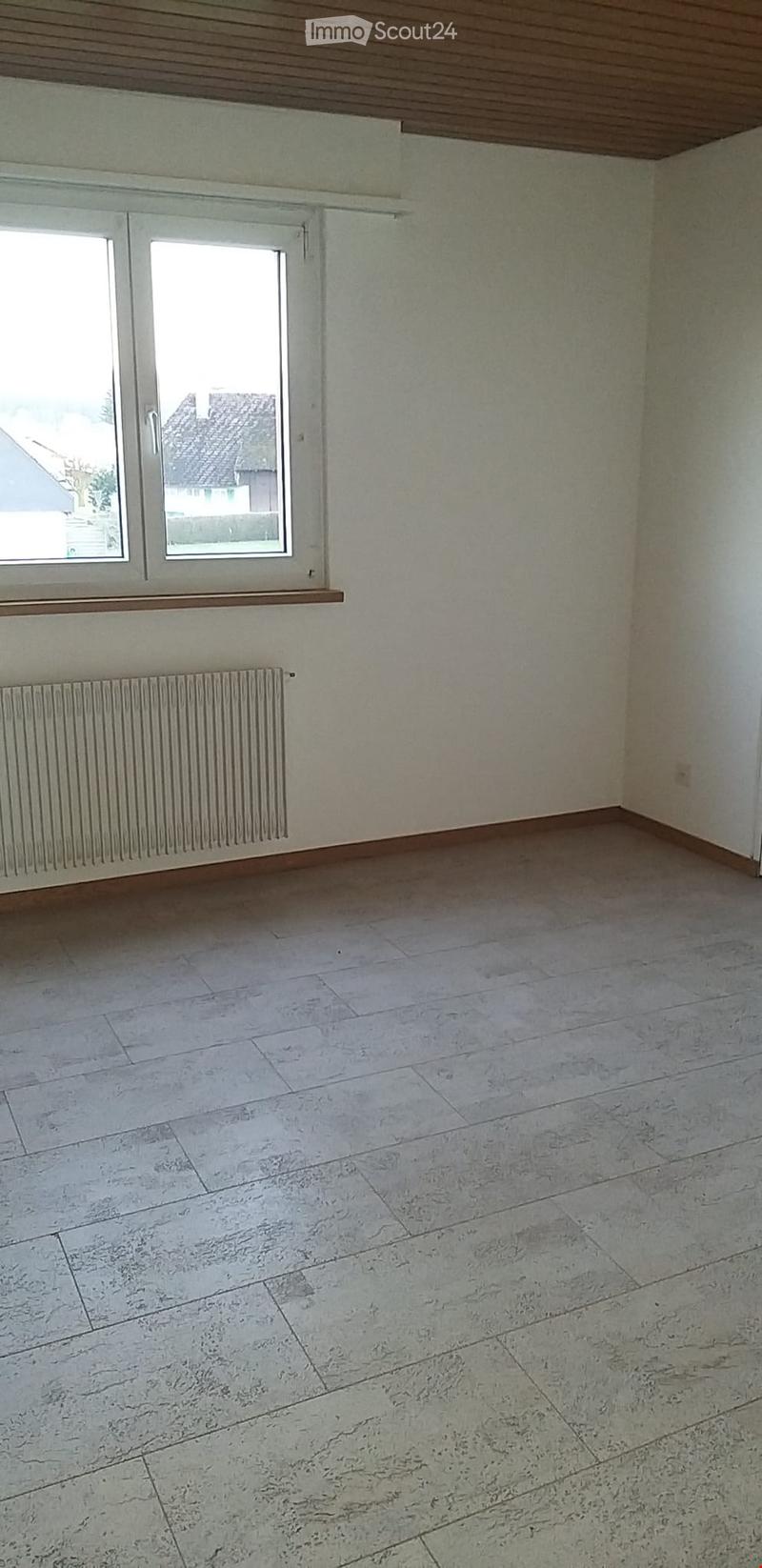 Empty room with light-colored walls, wooden ceiling, large window, radiator, and tiled floor.