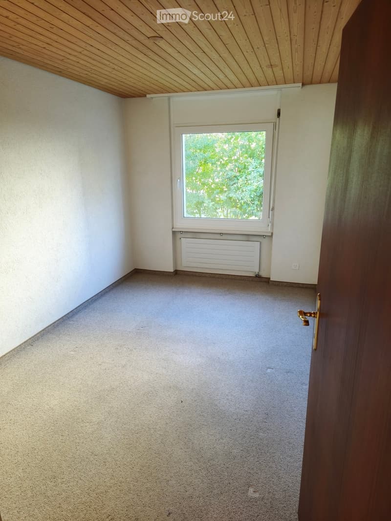 Empty room with light-colored walls, carpeted floor, and a window with a radiator below it. The room has a wooden door frame visible on the right side.
