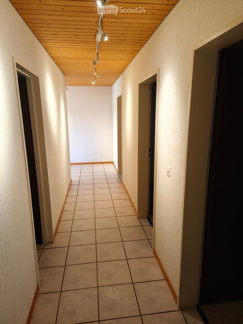 A narrow hallway with tiled flooring, light-colored walls, and a wooden ceiling with recessed lighting. Doors are visible on both sides.