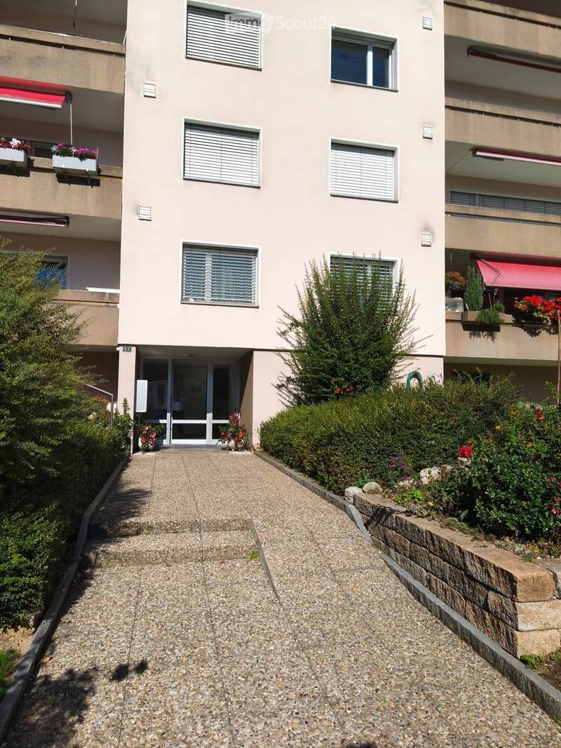 A multi-story residential building with beige walls, multiple windows with white blinds, and flower boxes on the balconies. The entrance features a glass door with a small garden area and a gravel pathway leading to it.