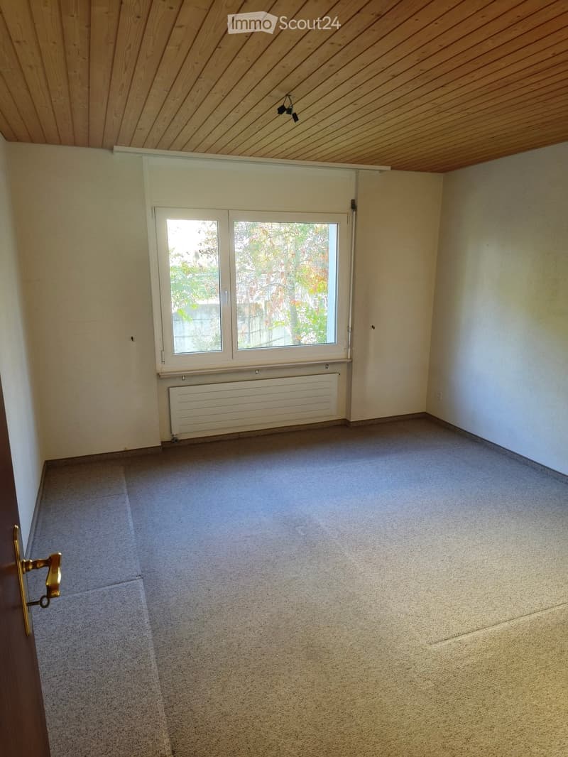 Empty room with light-colored walls, carpeted floor, and a large window with a radiator below it. The room has a wooden slanted ceiling and a door with a brass handle visible on the left side.