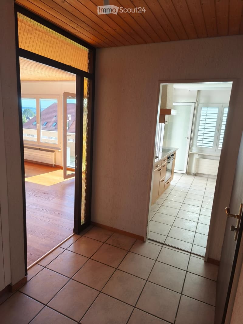 Hallway with tiled floor, leading to a kitchen area and an open space with wooden flooring and large windows offering a view of the outside.