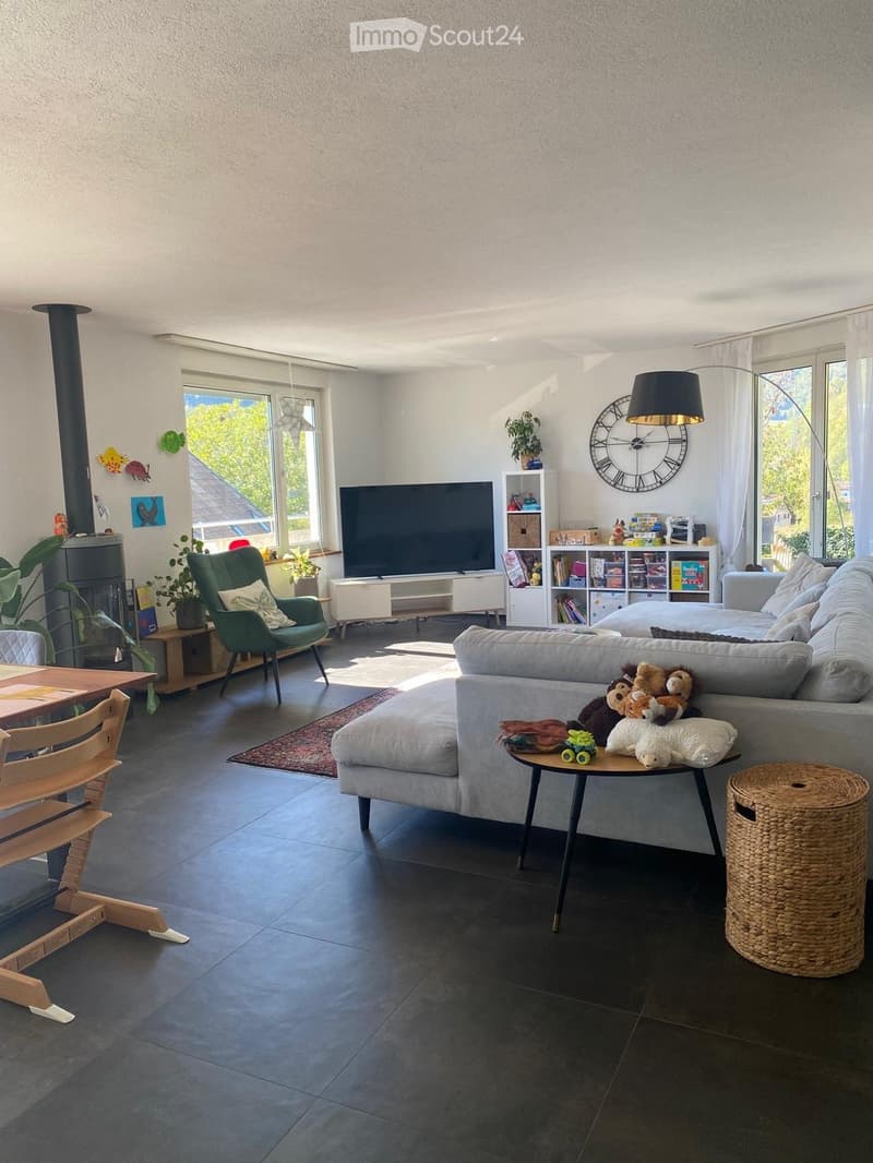 Modern living room with grey sectional sofa, coffee table, green armchair, TV, bookshelf, and fireplace. Natural light from large windows.