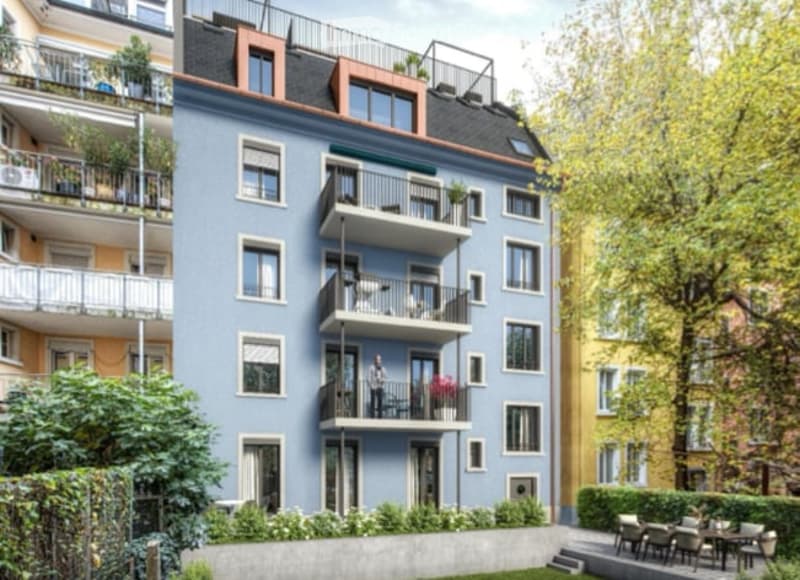 Modern multi-story apartment building with blue facade, multiple balconies, and greenery around the base.