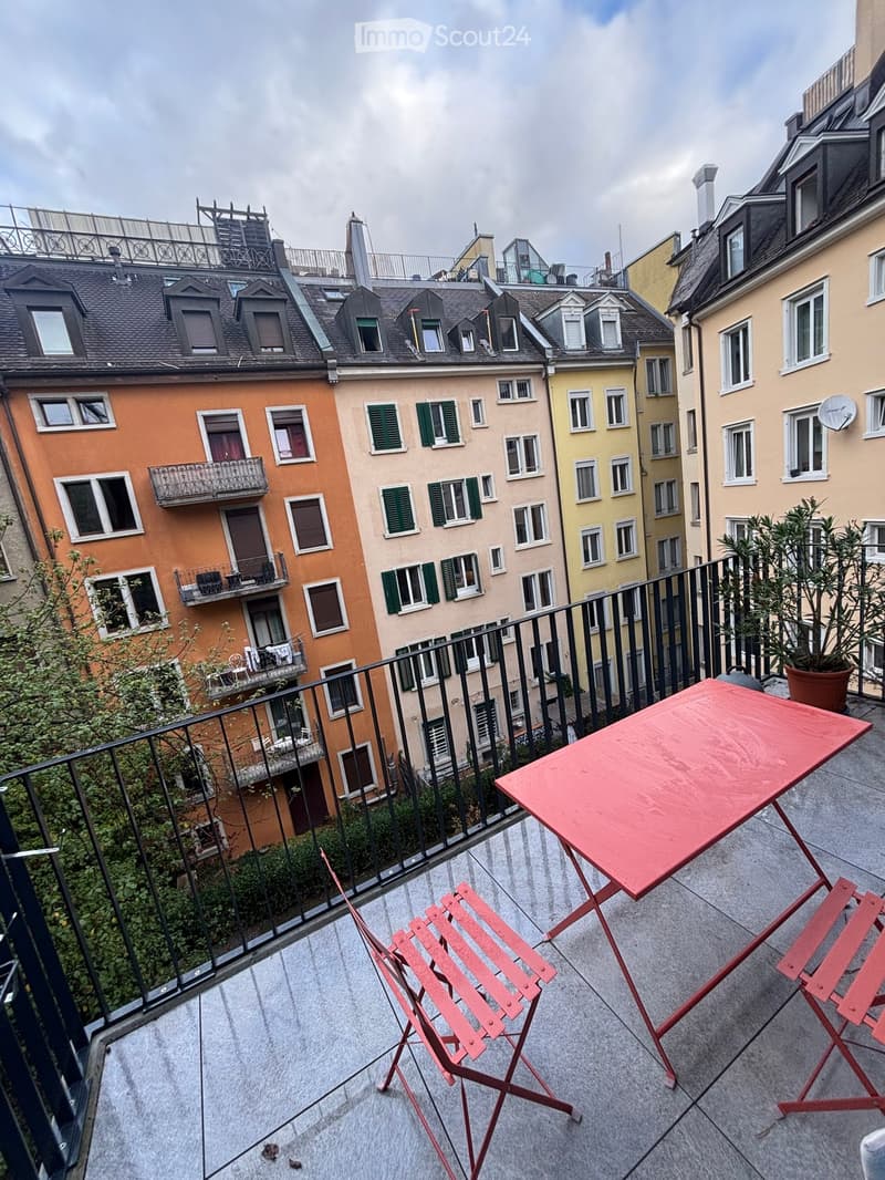 Balcony with a red table, two red chairs, and a potted plant, overlooking a cityscape with colorful buildings and multiple balconies.
