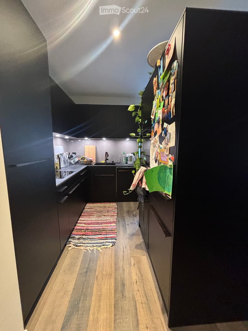 Modern kitchen with dark cabinets, wooden flooring, and a small rug. Features a refrigerator with magnets, a sink with a faucet, and a cutting board on the counter.
