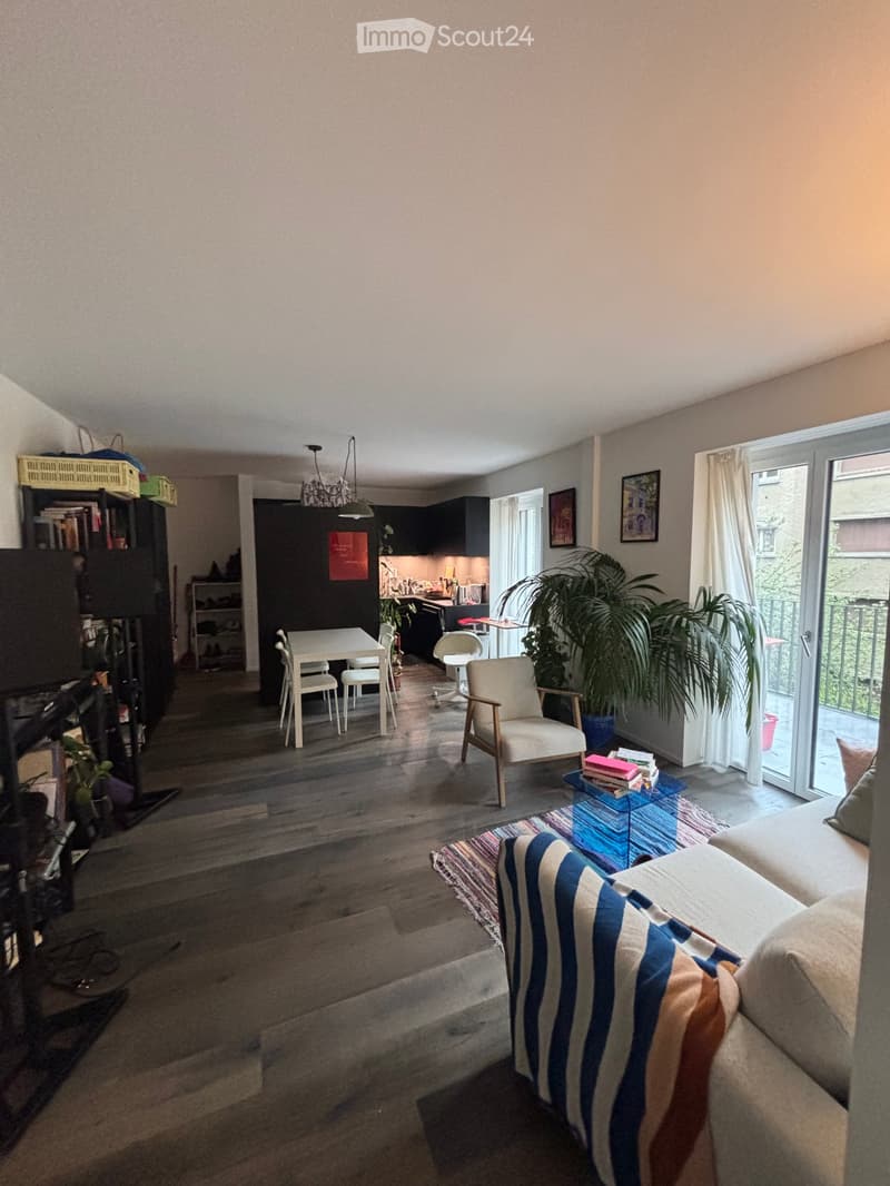 Open-plan living and dining area with wooden flooring, a sofa, armchair, coffee table, dining table with chairs, bookshelf, and a small kitchen area with cabinets and a stool.