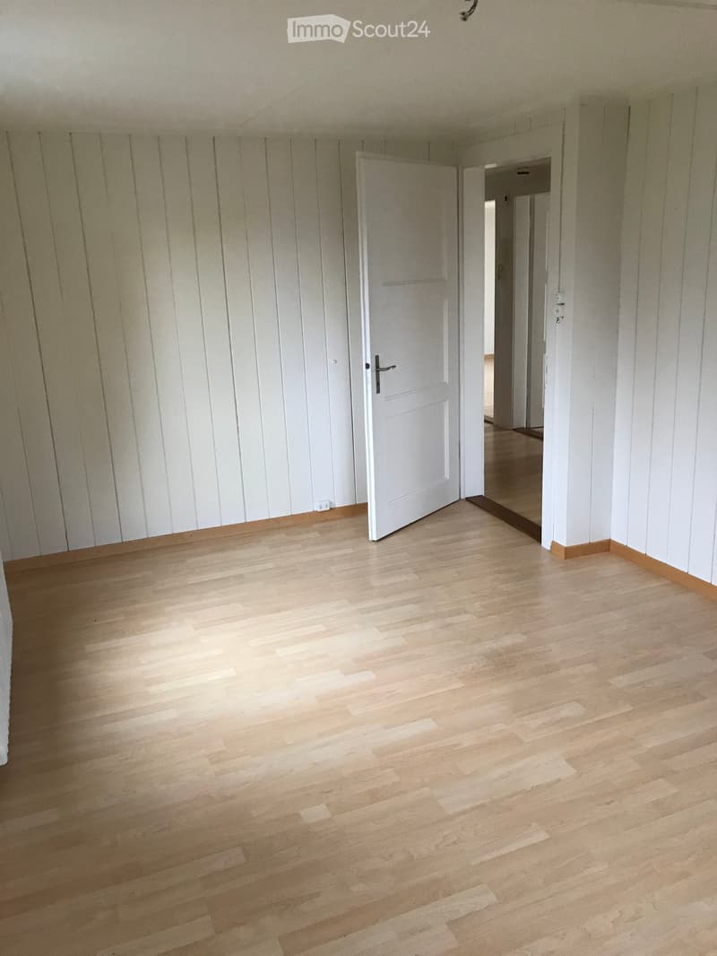Empty room with light wooden flooring and white paneled walls, featuring a white door and an open doorway leading to another space.