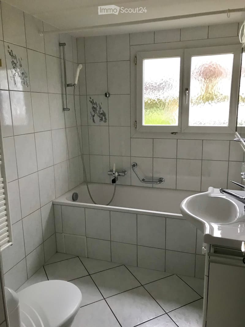 Bathroom with tiled walls and floor, featuring a bathtub, toilet, sink, and showerhead. There is a window with frosted glass and a handrail next to the bathtub.