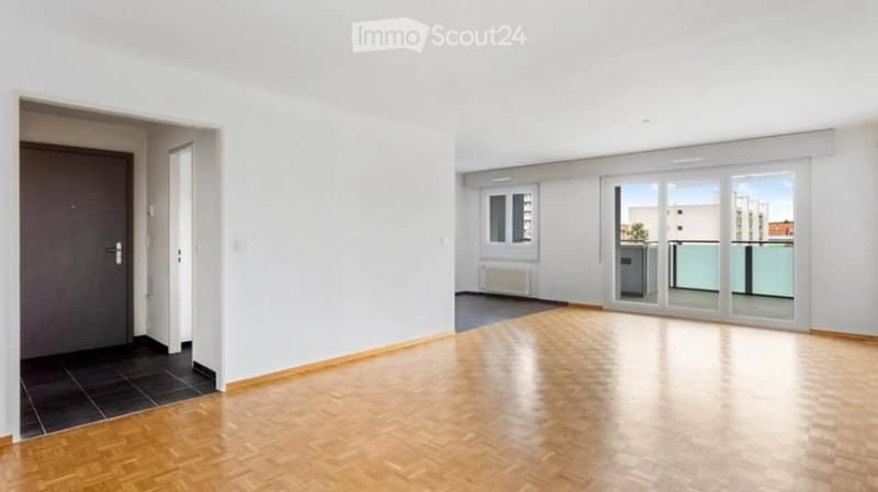 Empty room with wooden flooring, white walls, and large windows leading to a balcony area.