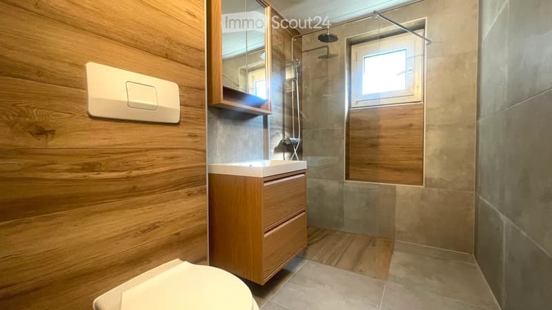 Modern bathroom with wooden walls, a white sink with a wooden cabinet, a toilet, a shower with a glass enclosure, and a small window for natural light.