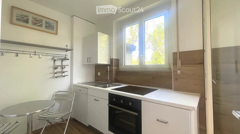 Modern kitchen with white cabinets, stainless steel appliances, a sink, stove, oven, and a small table with a chair. The space features wooden flooring and a large window with a view of greenery.