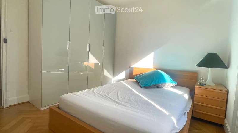 A modern bedroom with a wooden bed, white bedding, a blue pillow, a nightstand with a lamp, and a large mirrored wardrobe.