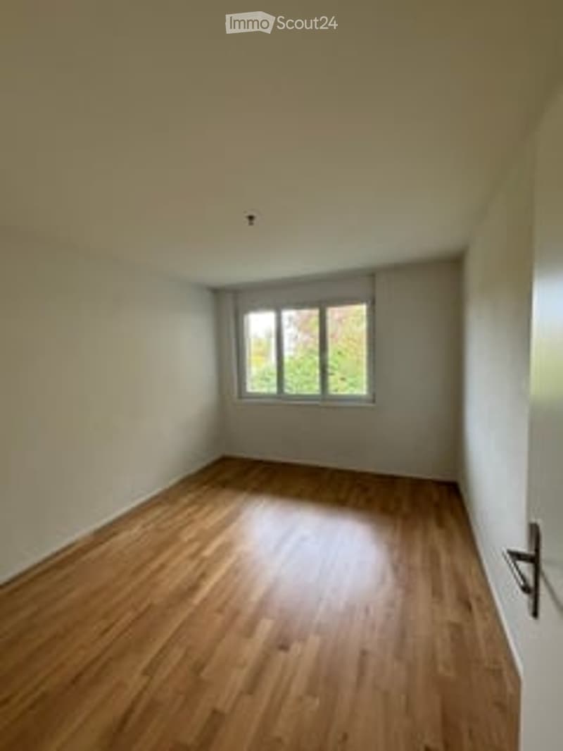 Completely empty room with wooden flooring, white walls, and a single window with curtains.