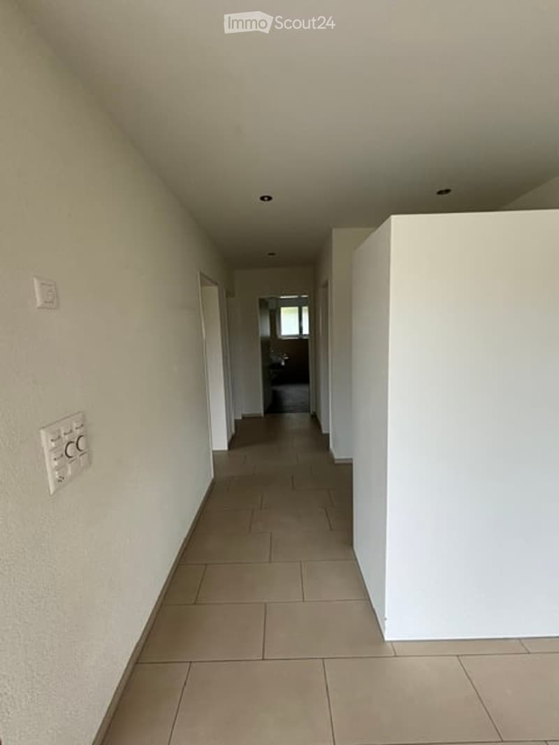 Empty hallway with beige tiled flooring, white walls, and recessed ceiling lighting. The corridor leads to an open area with a visible door at the end.