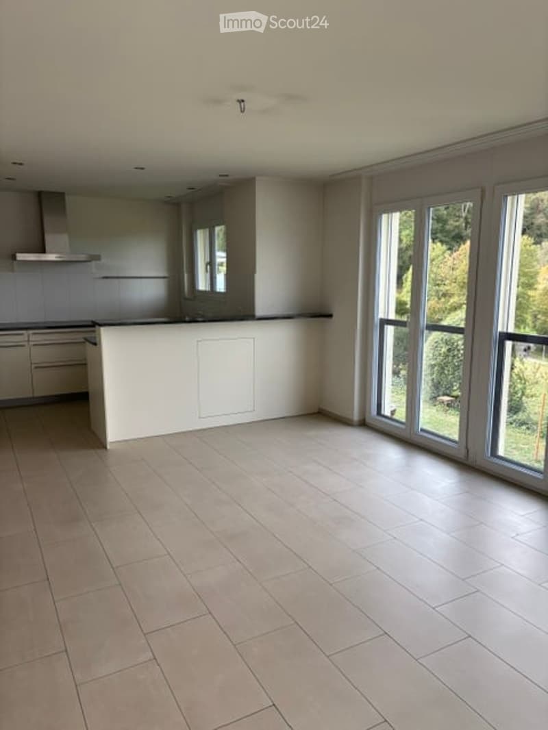 Open-plan space with a modern kitchen area and large windows leading to a garden. The floor is tiled, and the kitchen features light-colored cabinets and a central island.