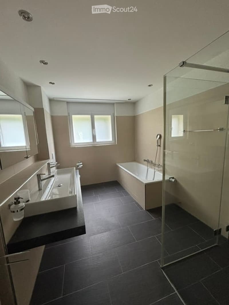 Modern bathroom with a bathtub, double sink, large mirror, and glass-enclosed shower. Dark tiled floor and light-colored walls.