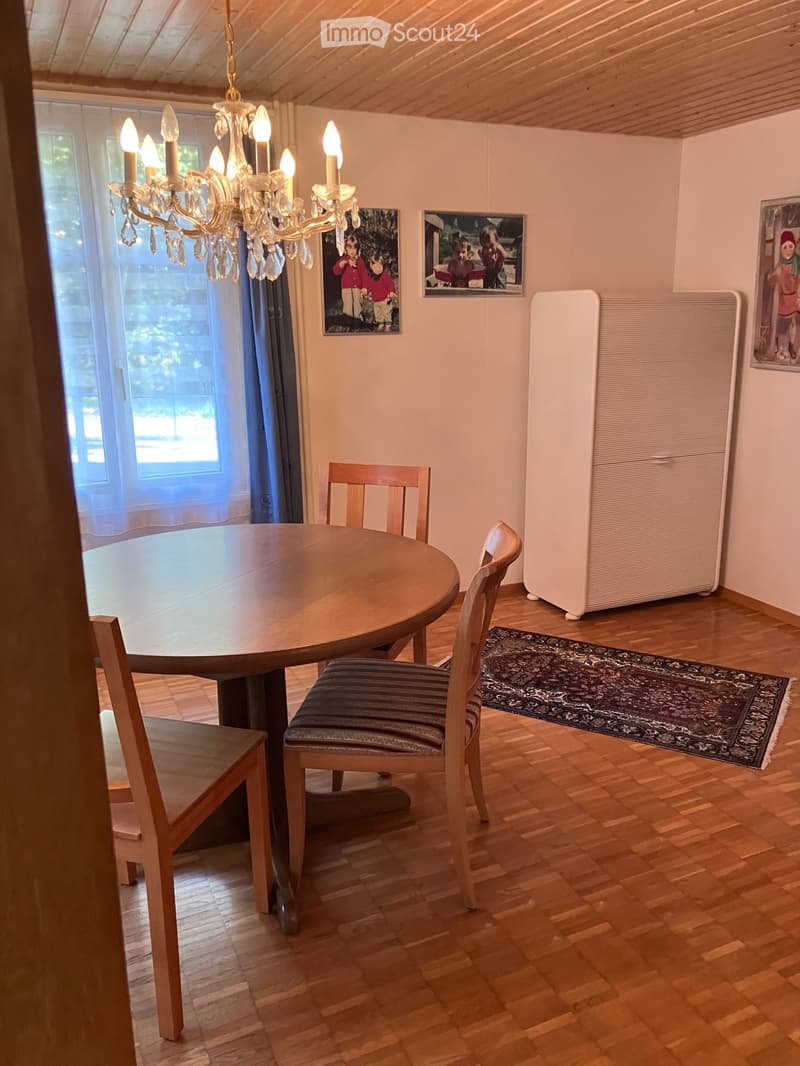 Bright kitchen with wooden flooring, round wooden table, four chairs, chandelier, radiator, and picture frames on the wall.