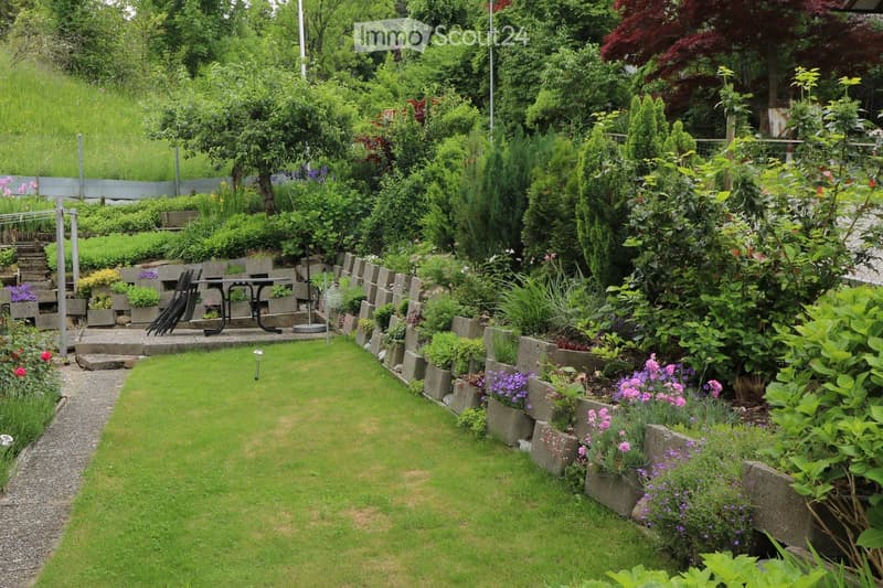 Well-maintained garden with a central lawn, raised stone planters, various flowering plants, shrubs, and a small seating area with a table and chairs.
