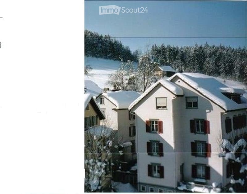 A multi-story residential building with snow-covered roofs and red-framed windows, surrounded by snow-covered trees and other houses in a winter landscape.
