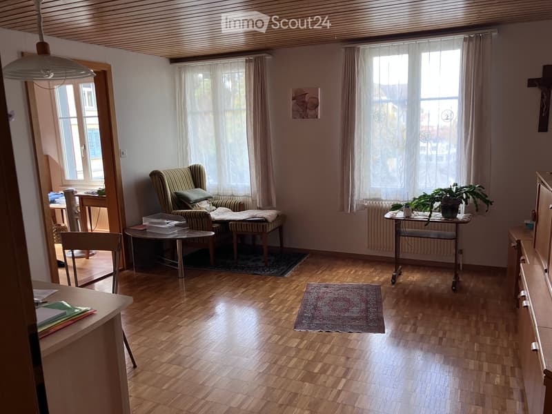 Living room with wooden flooring, large windows with curtains, a green armchair, a small table, a potted plant, and a cabinet.