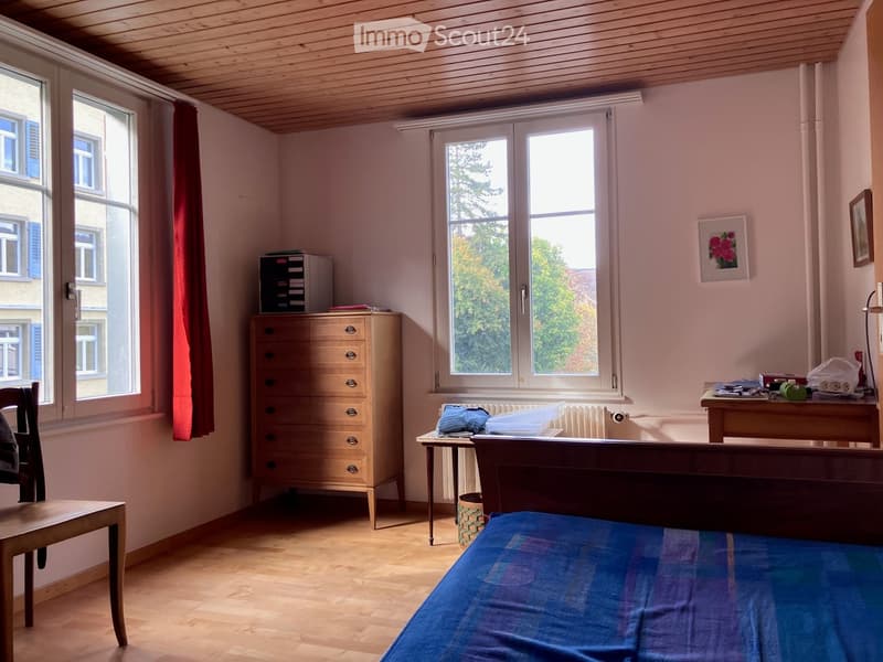 Cozy bedroom with wooden floor, large windows, bed with blue bedding, small table, chair, and dresser.