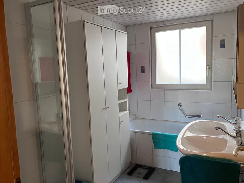Modern bathroom with a bathtub, sink, mirror, and white cabinet. Features a glass shower enclosure and a window with blinds.