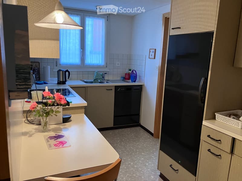 Modern kitchen with light wood cabinets, stainless steel appliances, a central island with a vase of pink flowers, and a hanging light fixture.
